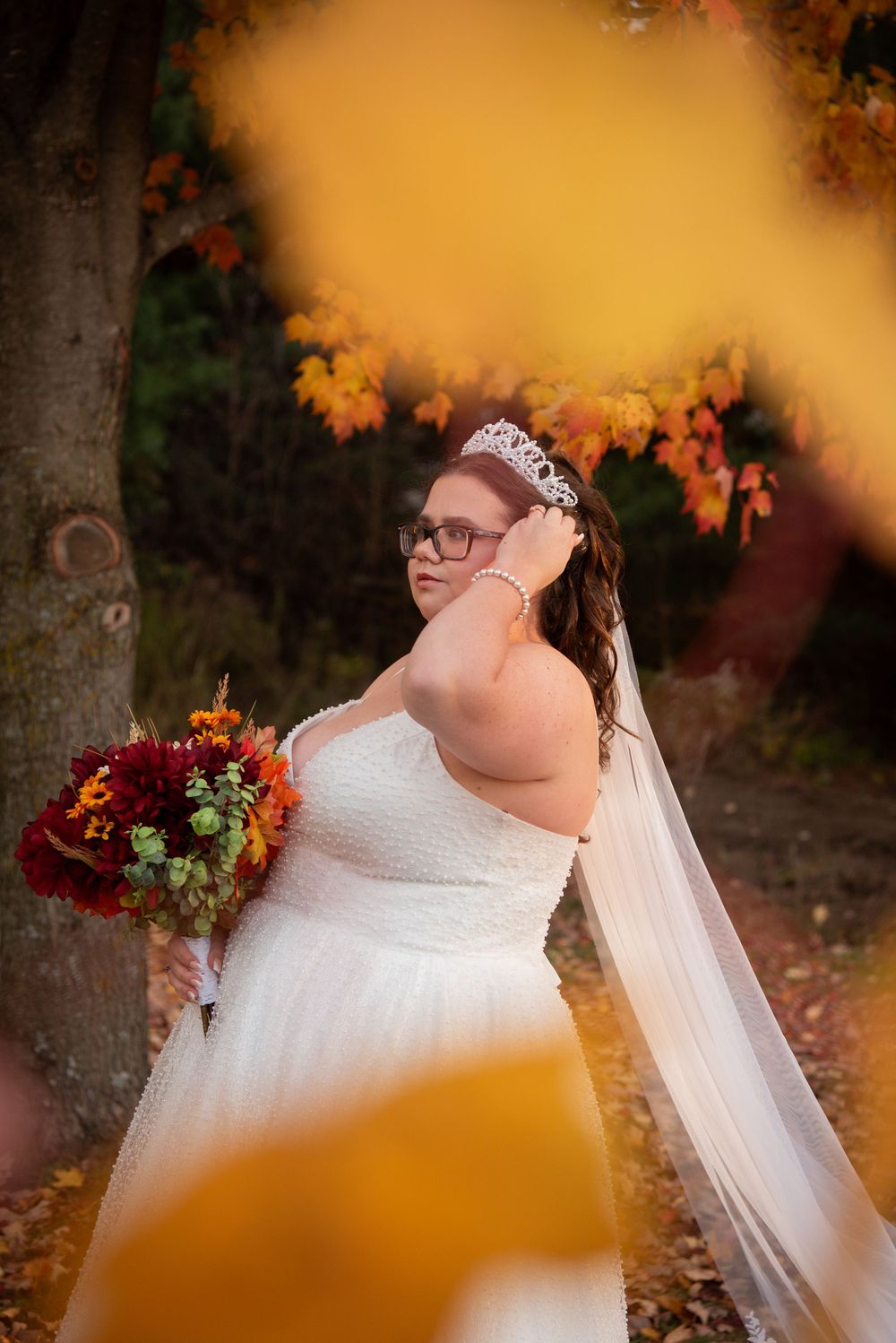A bride in a white dress and veil poses outdoors against autumn foliage with orange bokeh effects in the foreground.