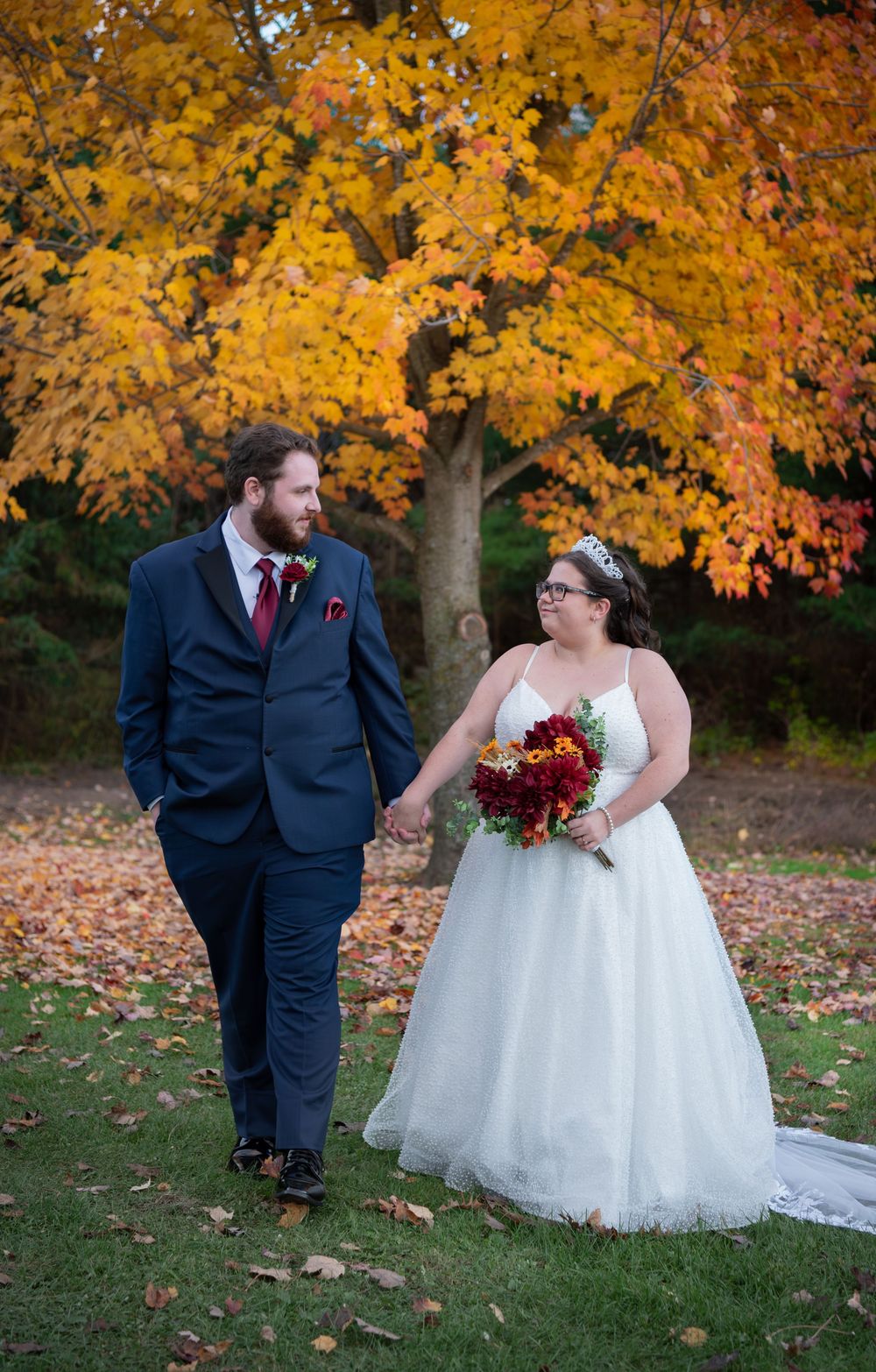 A couple in wedding attire stands together under vibrant golden autumn foliage in an outdoor setting.