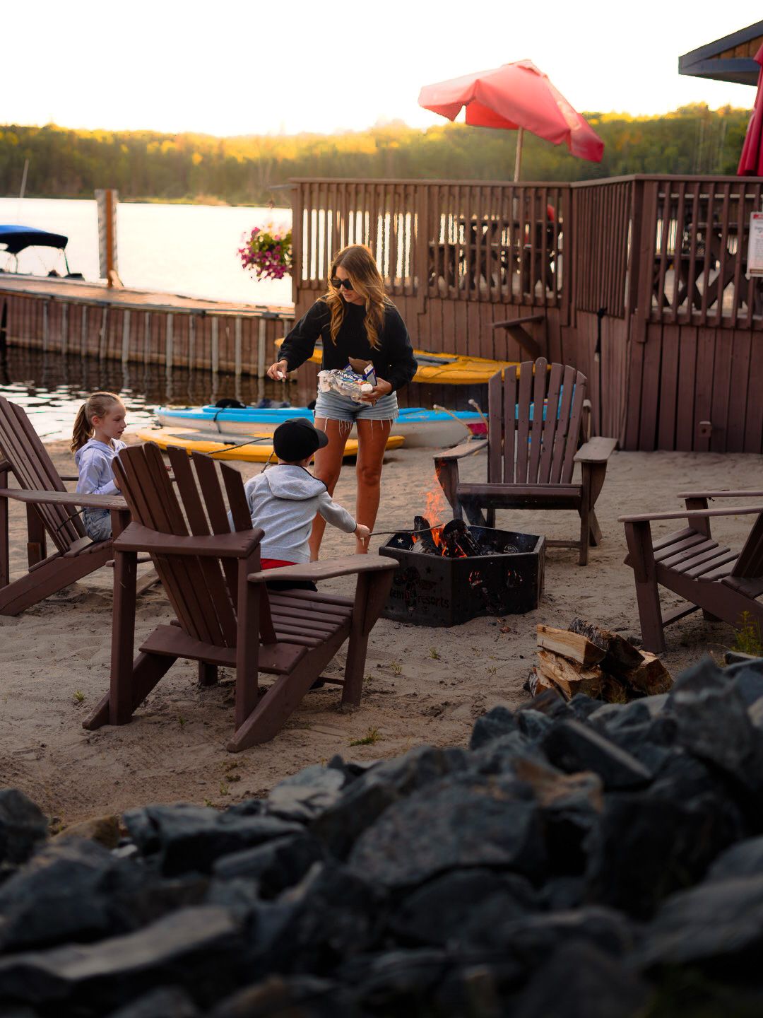 Wooden Adirondack chairs arranged on a beach at sunset with fire pit in foreground.