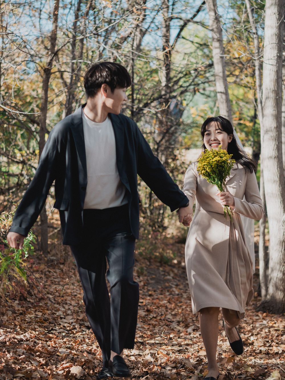 A couple playfully running through an autumn forest with fallen leaves scattered on the ground.