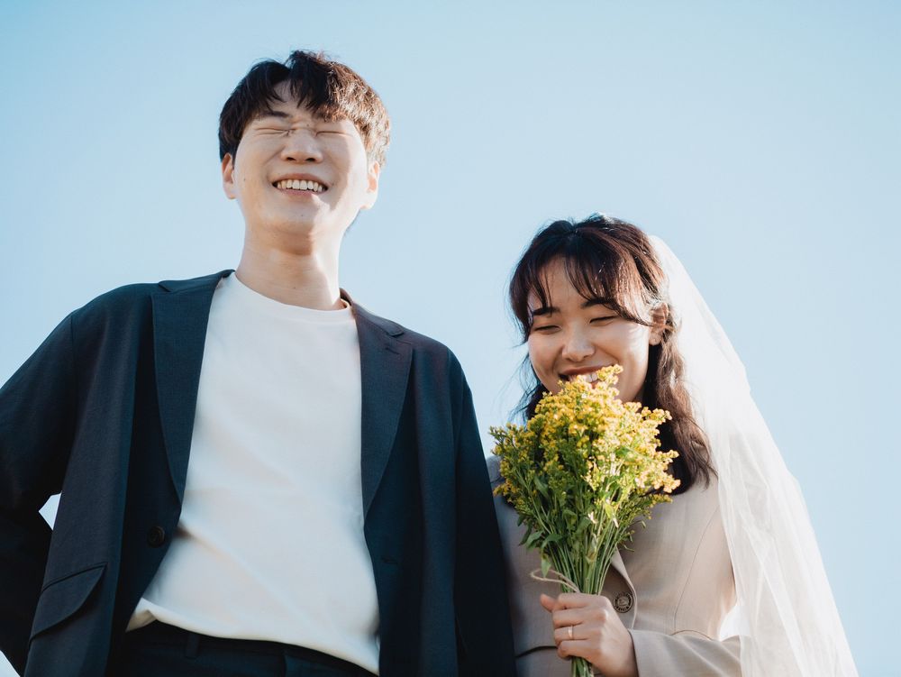 A couple in wedding attire shares joyful moments together while holding yellow flowers against a bright sky background.