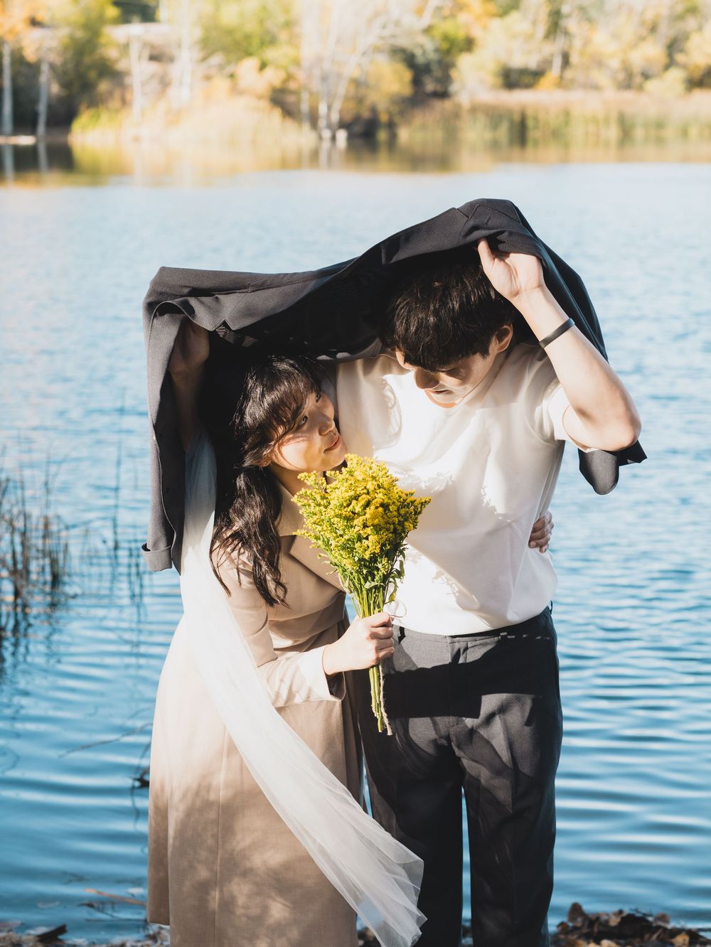 Romantic couple shares tender moments by a serene lake with yellow flowers and flowing veil in autumn light.