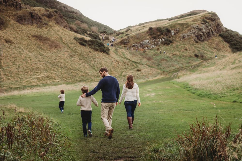 A family walks along a scenic mountain trail through rugged terrain and green hills on a cloudy day.