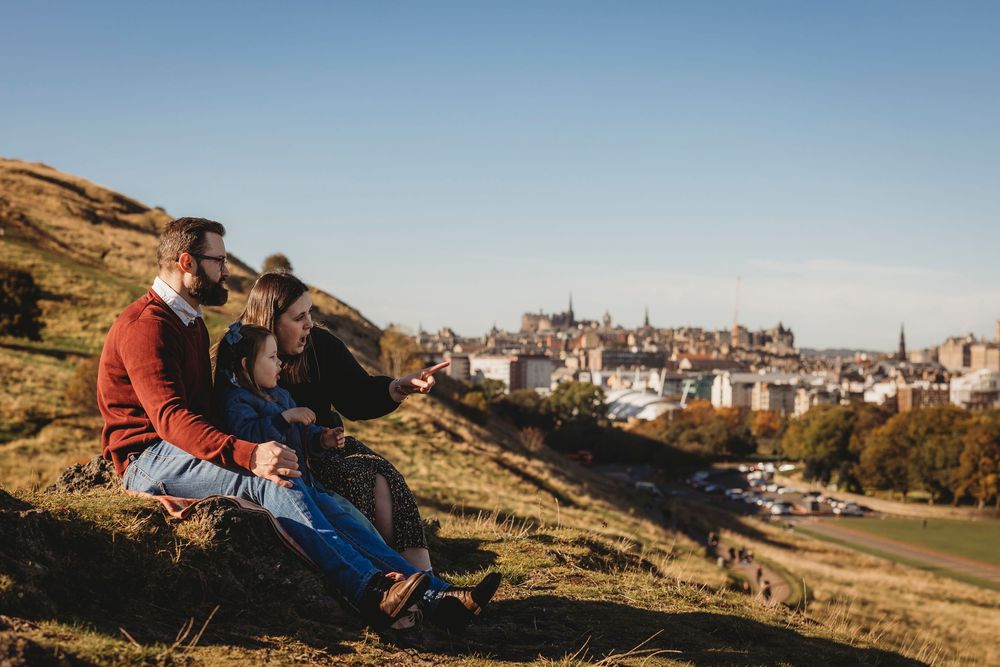 Two people sit together on a grassy hillside overlooking Edinburgh's historic skyline at sunset.