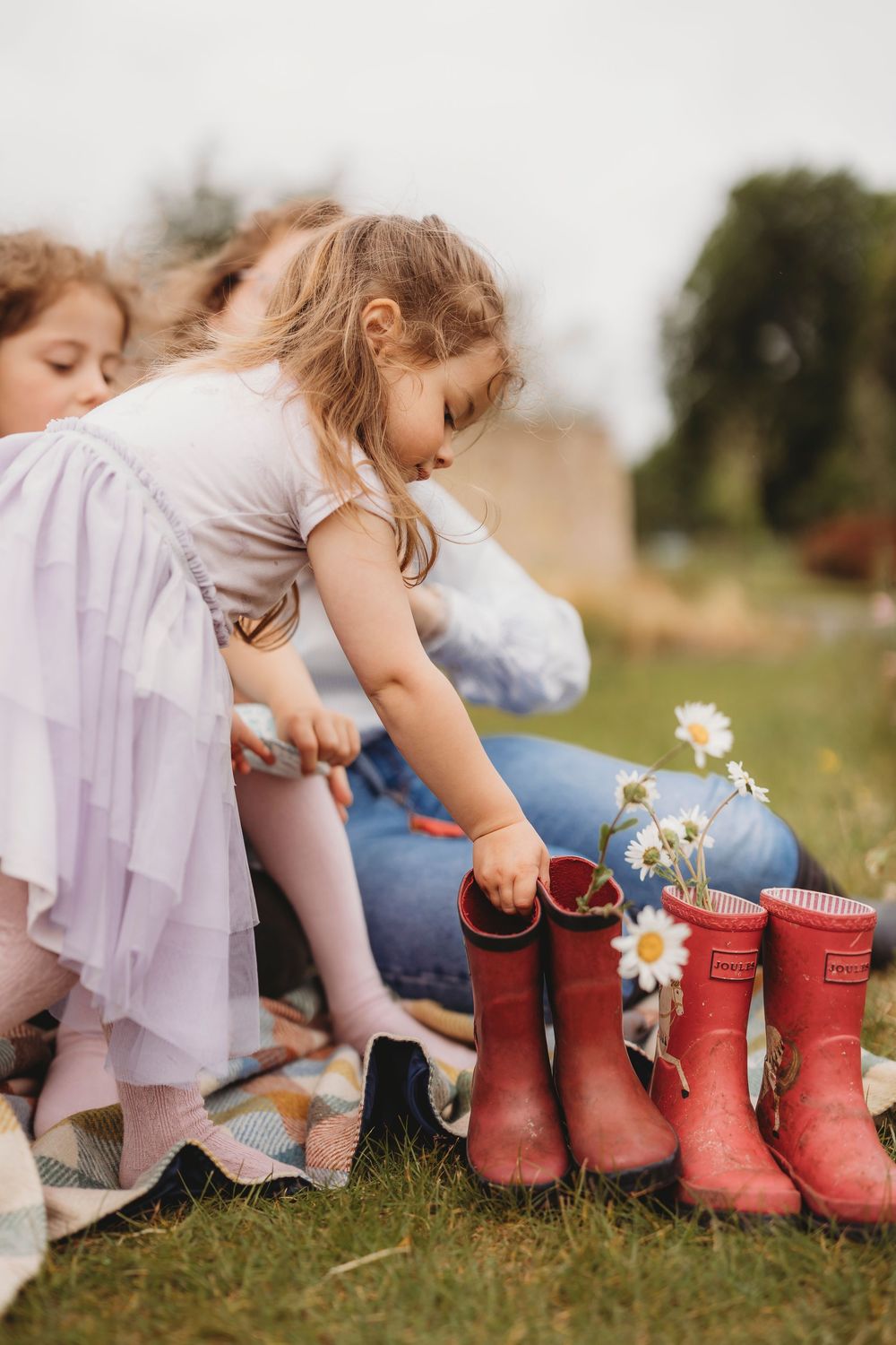 Series of red rain boots with daisies placed in them sitting on grass in a dreamy outdoor setting.