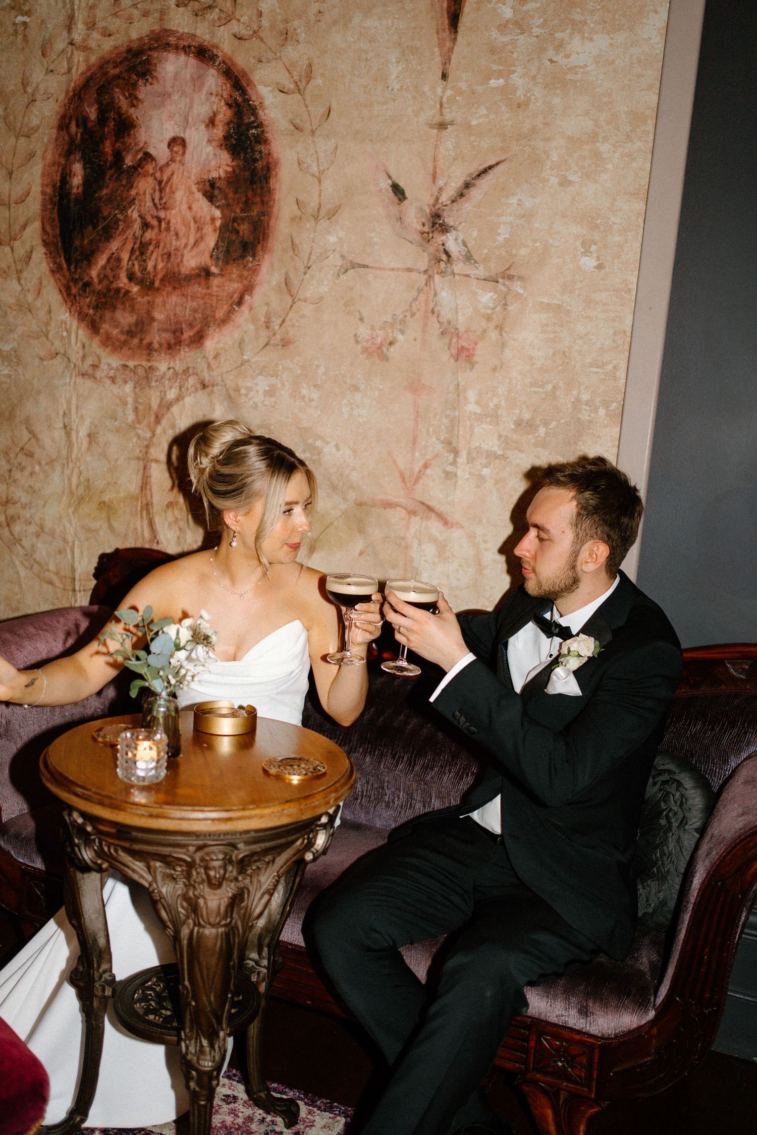A couple in wedding attire shares an intimate toast at a small table in a dimly lit vintage-style venue.