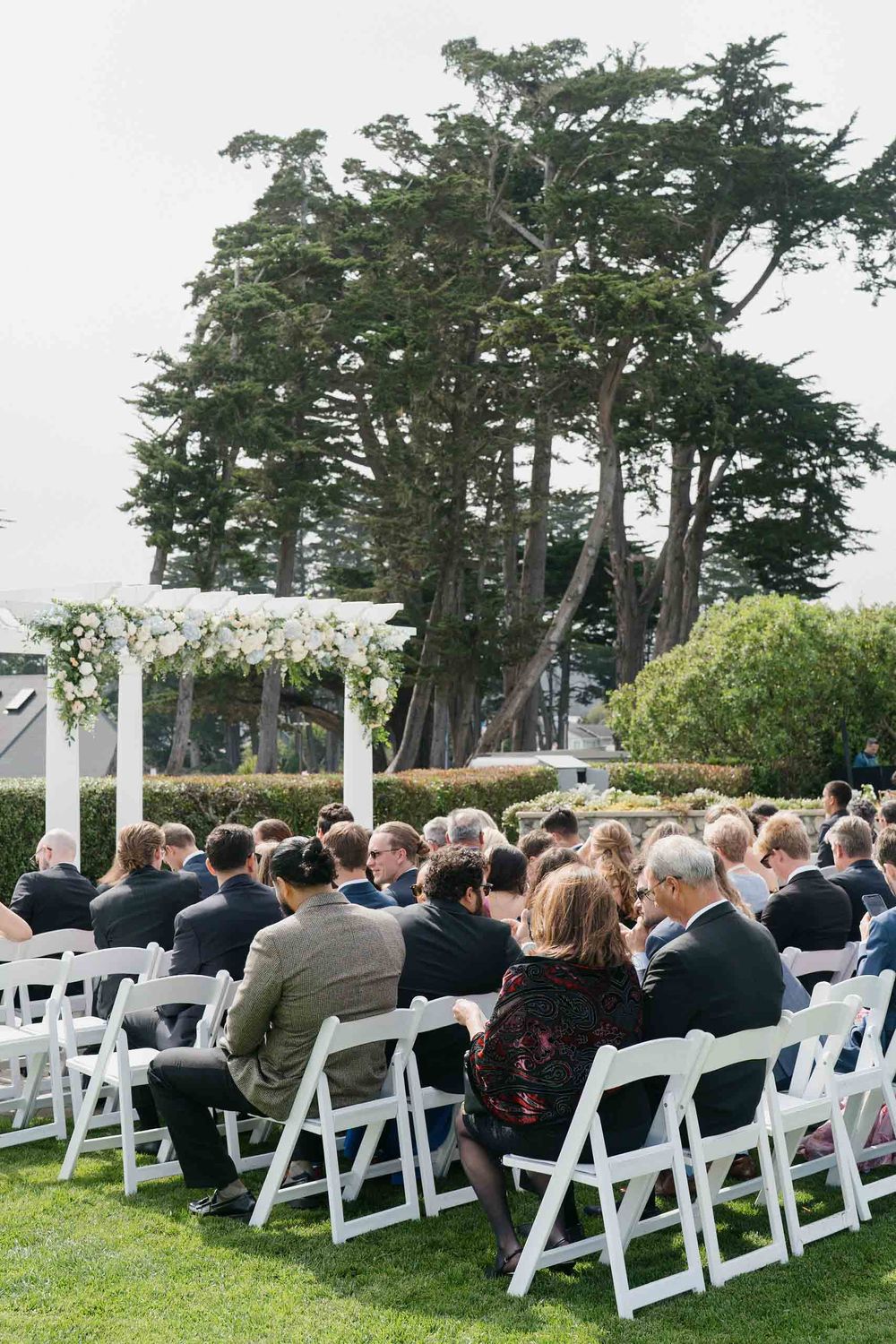 Wedding guests seated in white chairs on a lawn surrounded by tall trees during an outdoor ceremony.