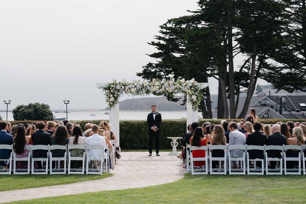 Outdoor wedding ceremony setup with white chairs arranged in rows under tall evergreen trees.