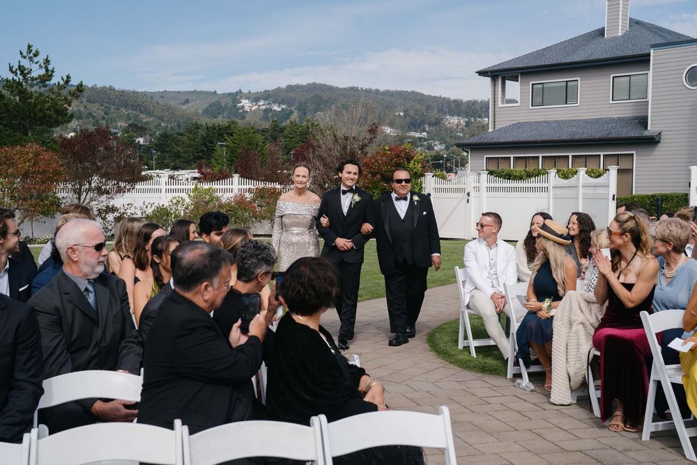 Wedding guests gather outdoors at a scenic venue with mountains and water views in the background.
