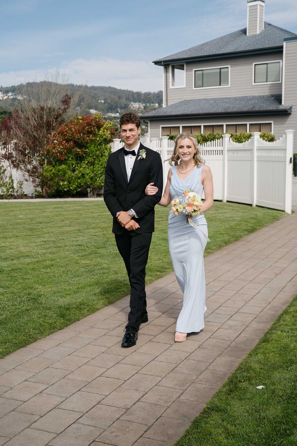 A couple in formal wedding attire walks along a paved pathway beside a green lawn and house.