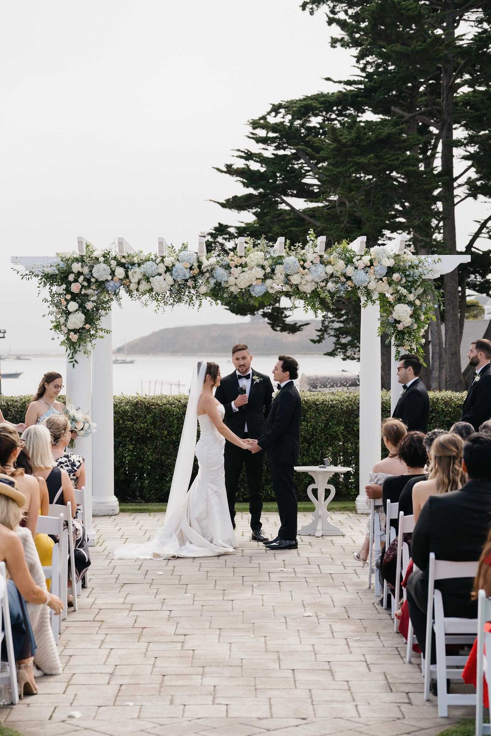A wedding ceremony under a white floral arch with guests seated on either side of a stone pathway.
