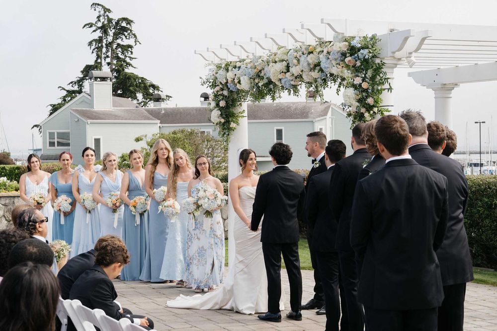 Wedding party members in blue dresses and black suits gather outdoors for a formal group photo.