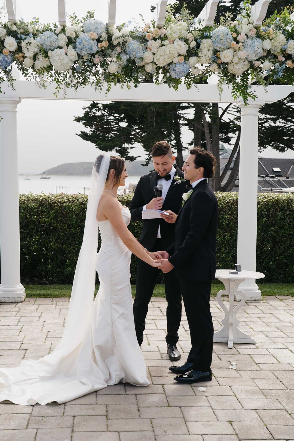 Wedding party gathers under floral arch on stone patio during outdoor ceremony with white columns and greenery.