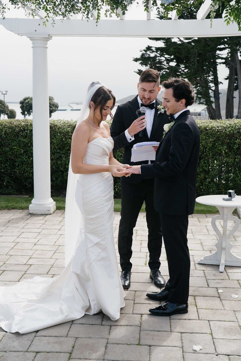 A wedding ceremony takes place outdoors on a stone patio with white columns and greenery overhead.