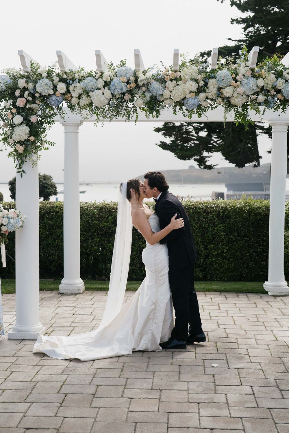 Romantic wedding ceremony sequence under a white pergola decorated with elegant floral arrangements and greenery.