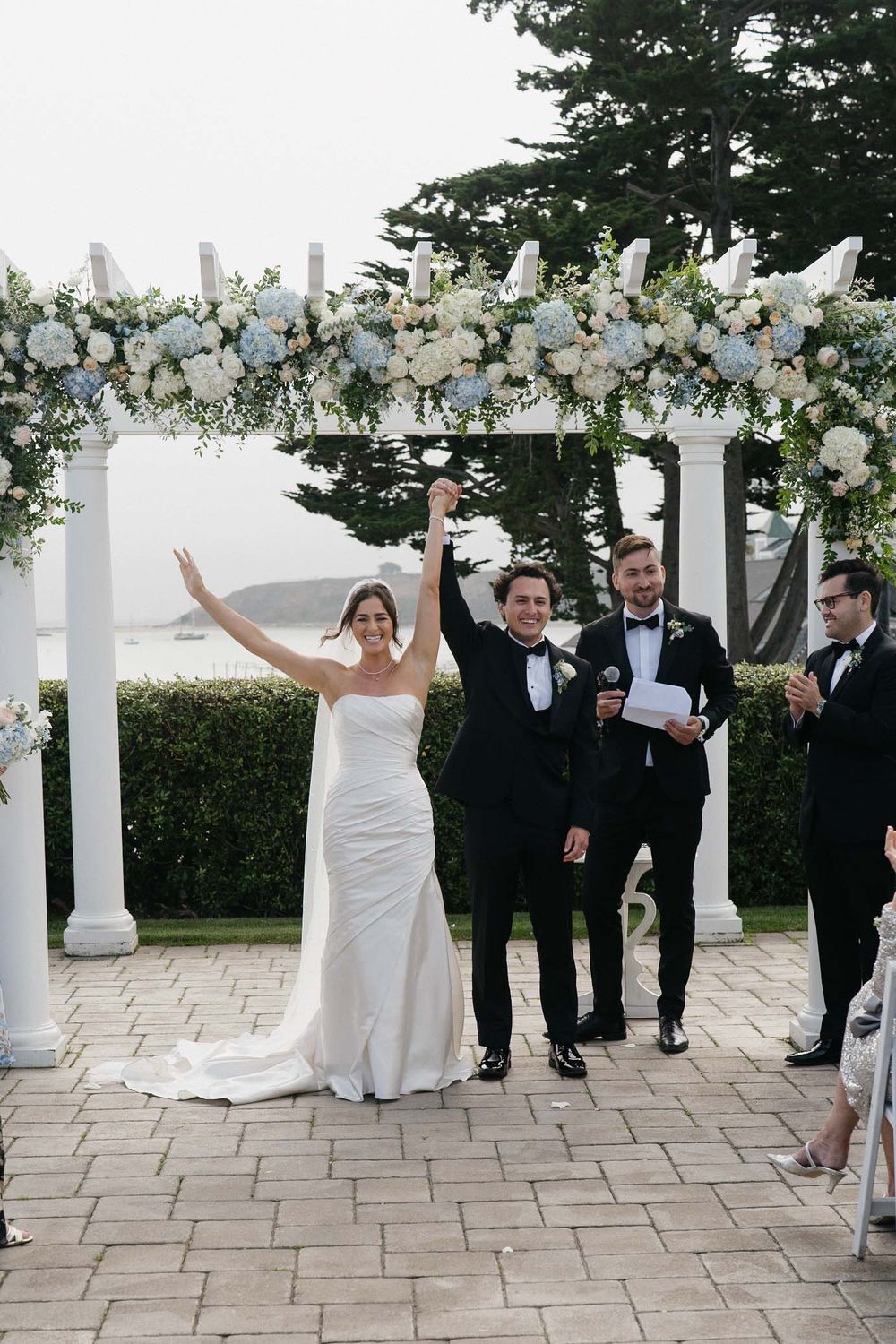 Newlyweds celebrate under an elegant white floral arch during an outdoor wedding ceremony.