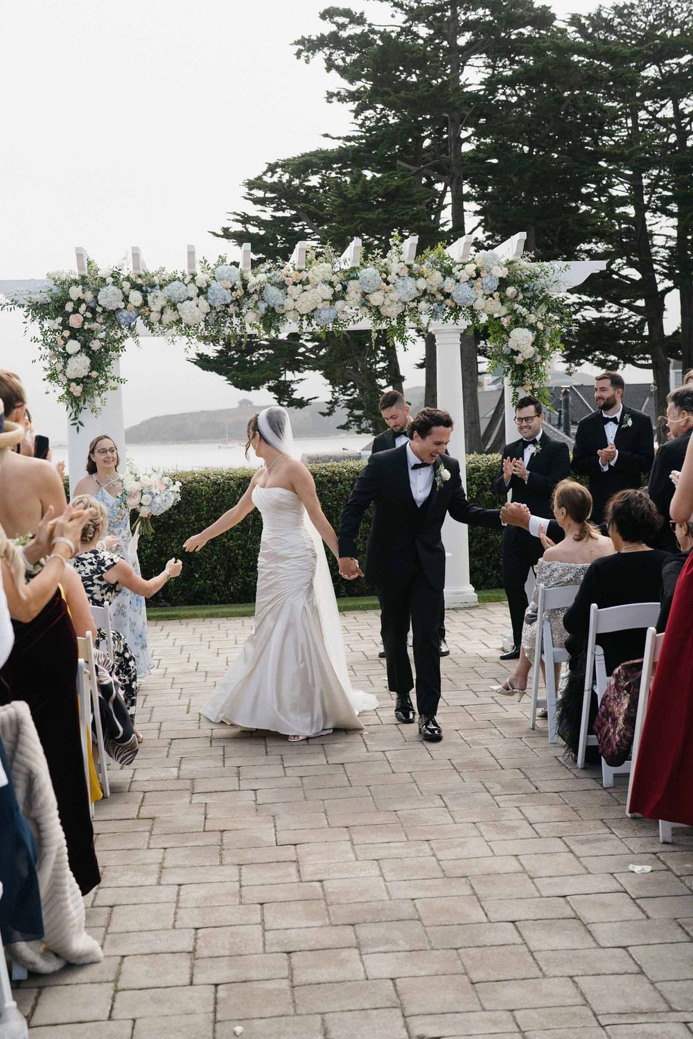Wedding guests applaud as the couple walks down the aisle under a beautiful white floral archway.