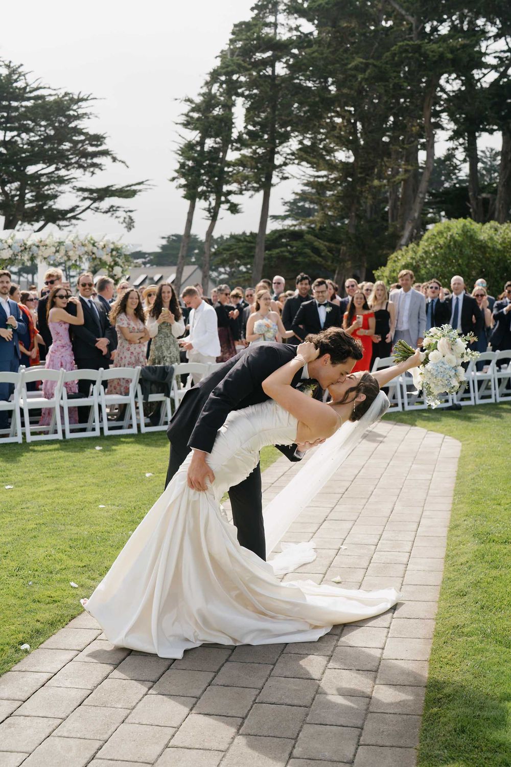 A romantic dip kiss on a stone pathway during an outdoor wedding celebration.