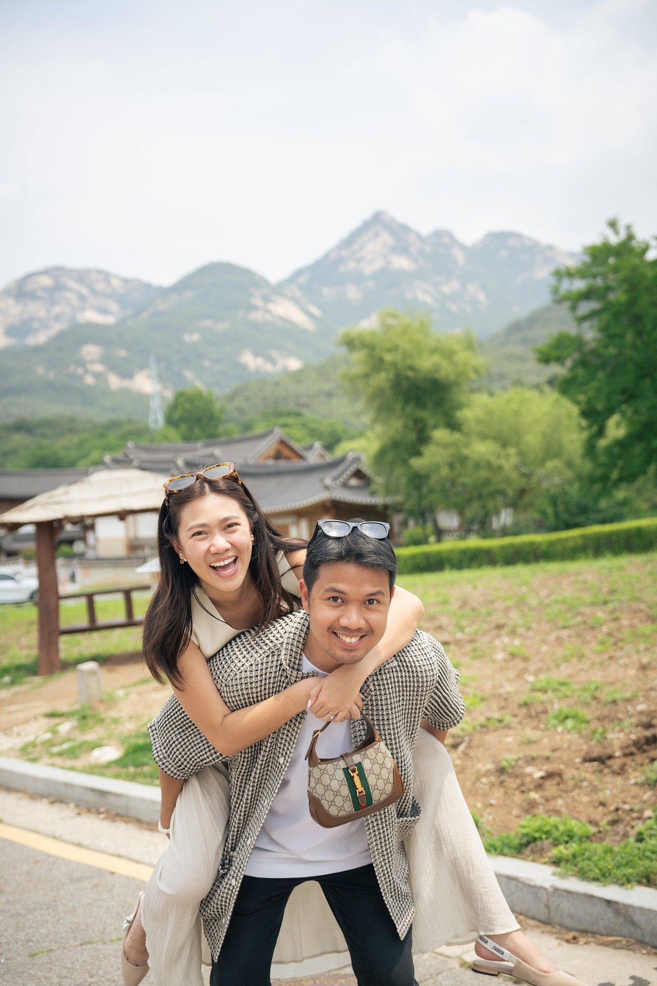 Two friends share a playful piggyback moment on a path with scenic mountains and traditional buildings in the background.