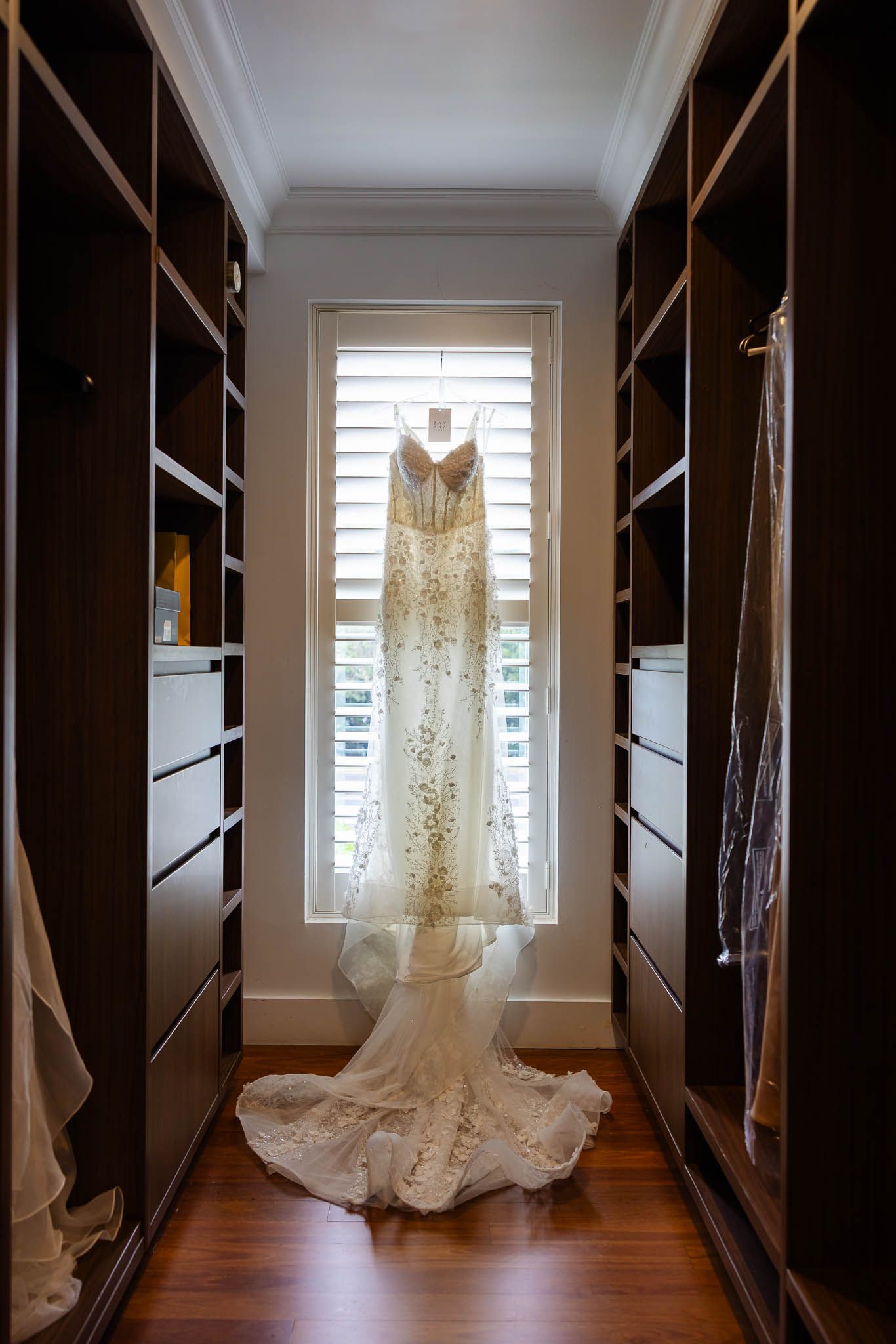 A wedding dress hangs elegantly in a walk-in closet with dark wood built-ins and natural light streaming through windows.