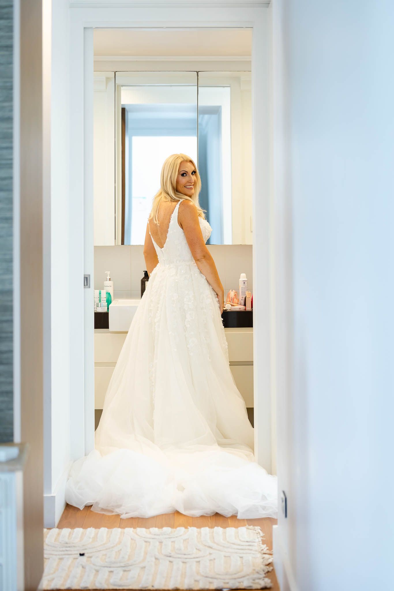 A bride in a flowing white wedding gown stands in a bright room with multiple mirrors creating an elegant reflection.