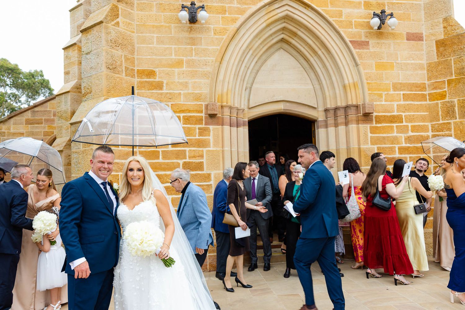 Wedding guests gather outside a beautiful sandstone church with gothic arched doorways and windows.