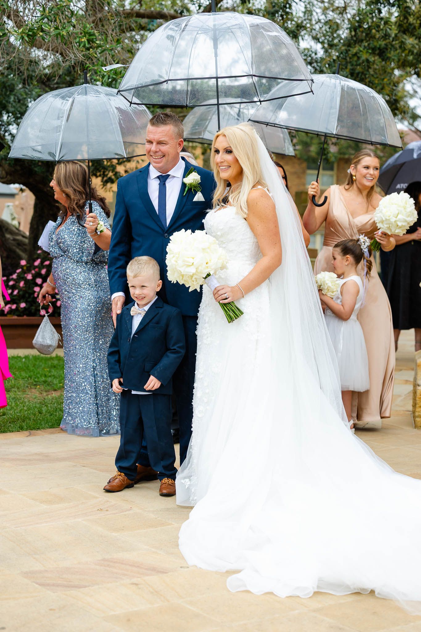 Wedding party stands under clear umbrellas on a rainy day during outdoor ceremony on concrete patio.