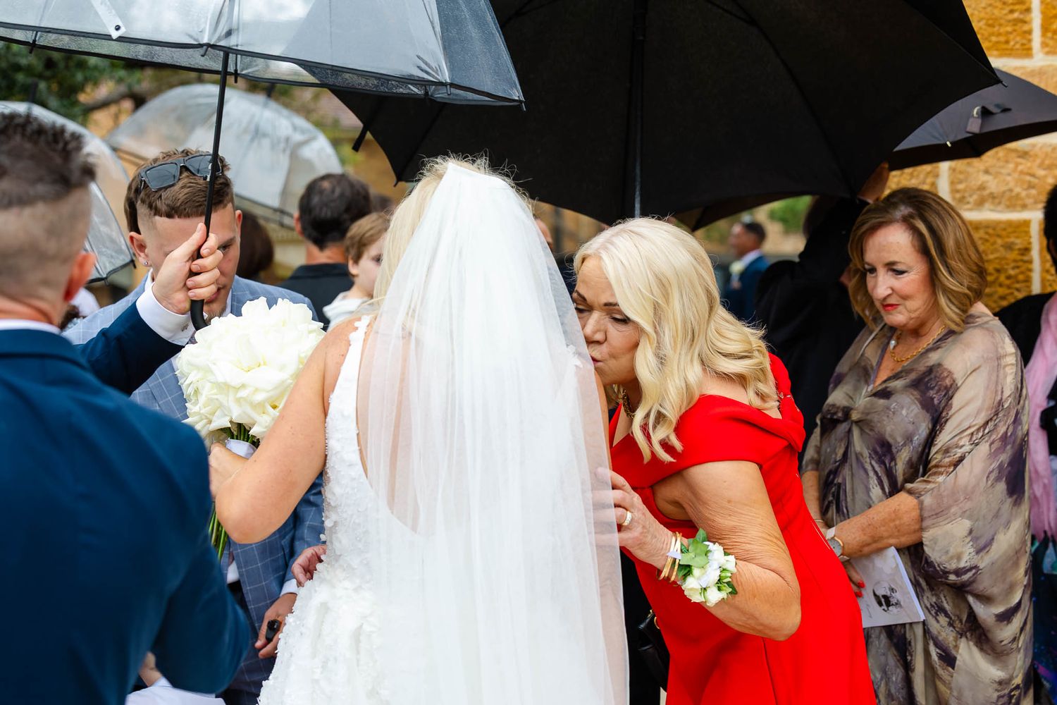A group of wedding guests huddle together under umbrellas during a rainy outdoor celebration.