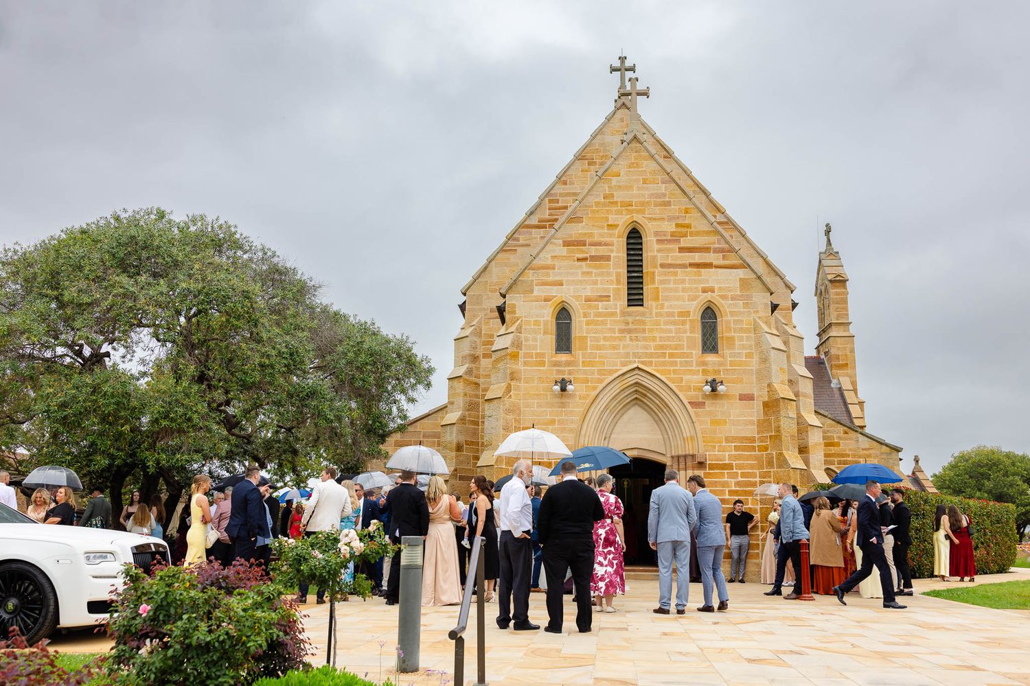 A stone church with a cross on top stands against a cloudy sky as people gather outside for a wedding ceremony.