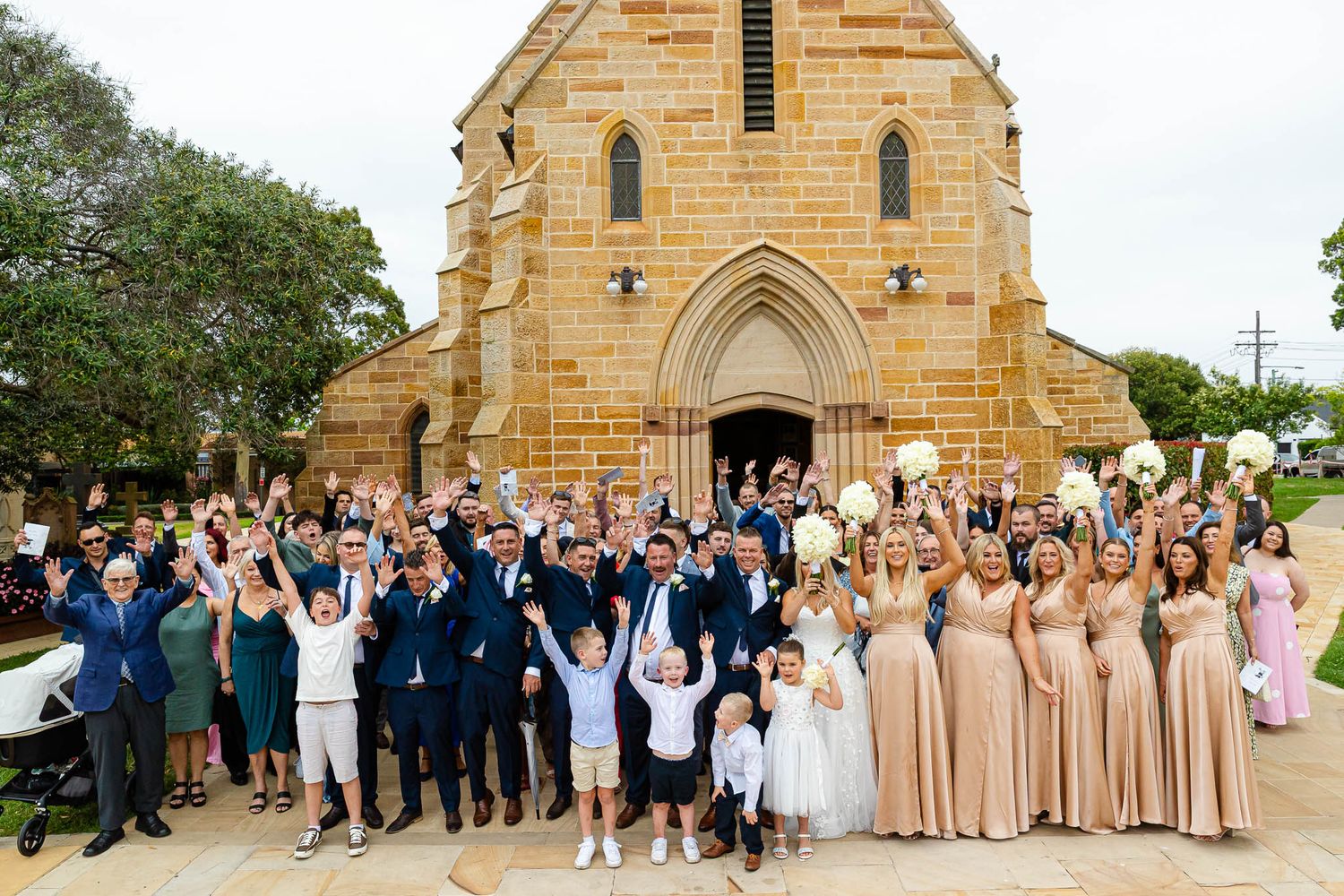 A large group celebrates outside a historic sandstone church with Gothic arched windows and doorway.