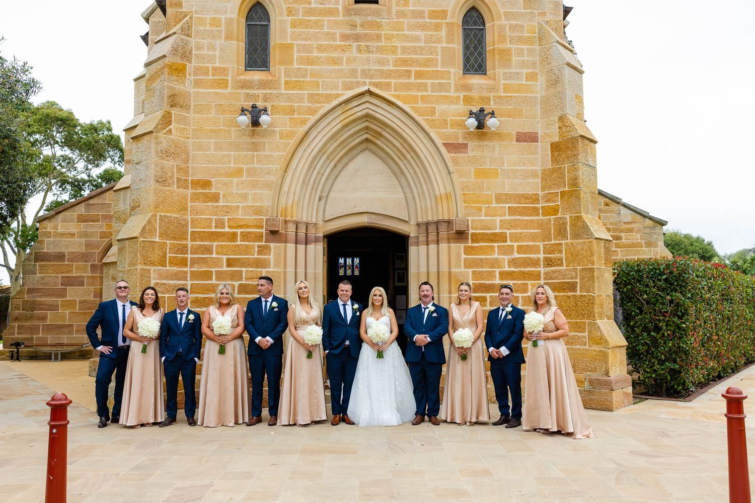Wedding party stands outside historic sandstone church with gothic arched doorway in panoramic view.