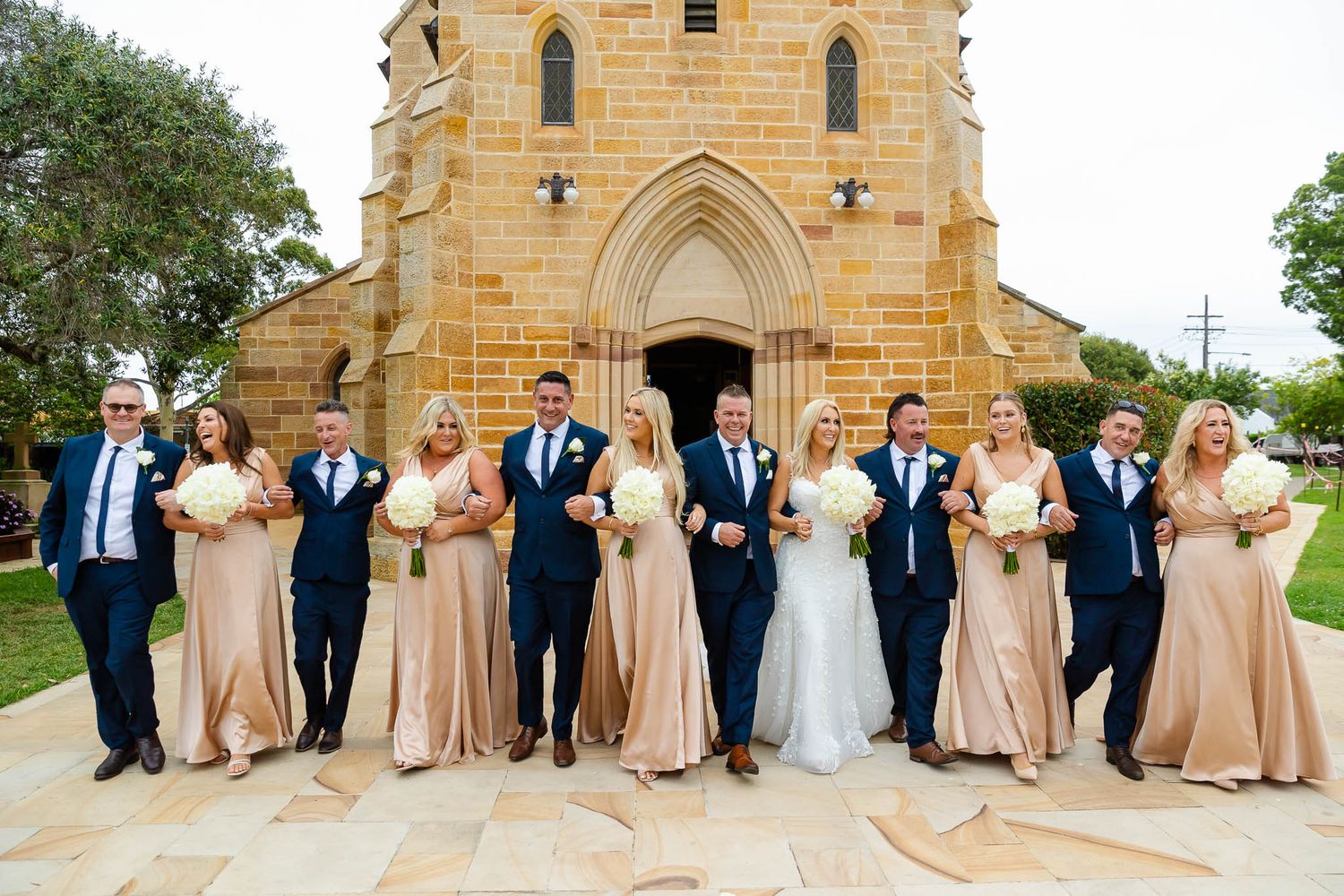 A wedding party in navy suits and champagne gowns poses in front of a stone church with Gothic arched entrance.