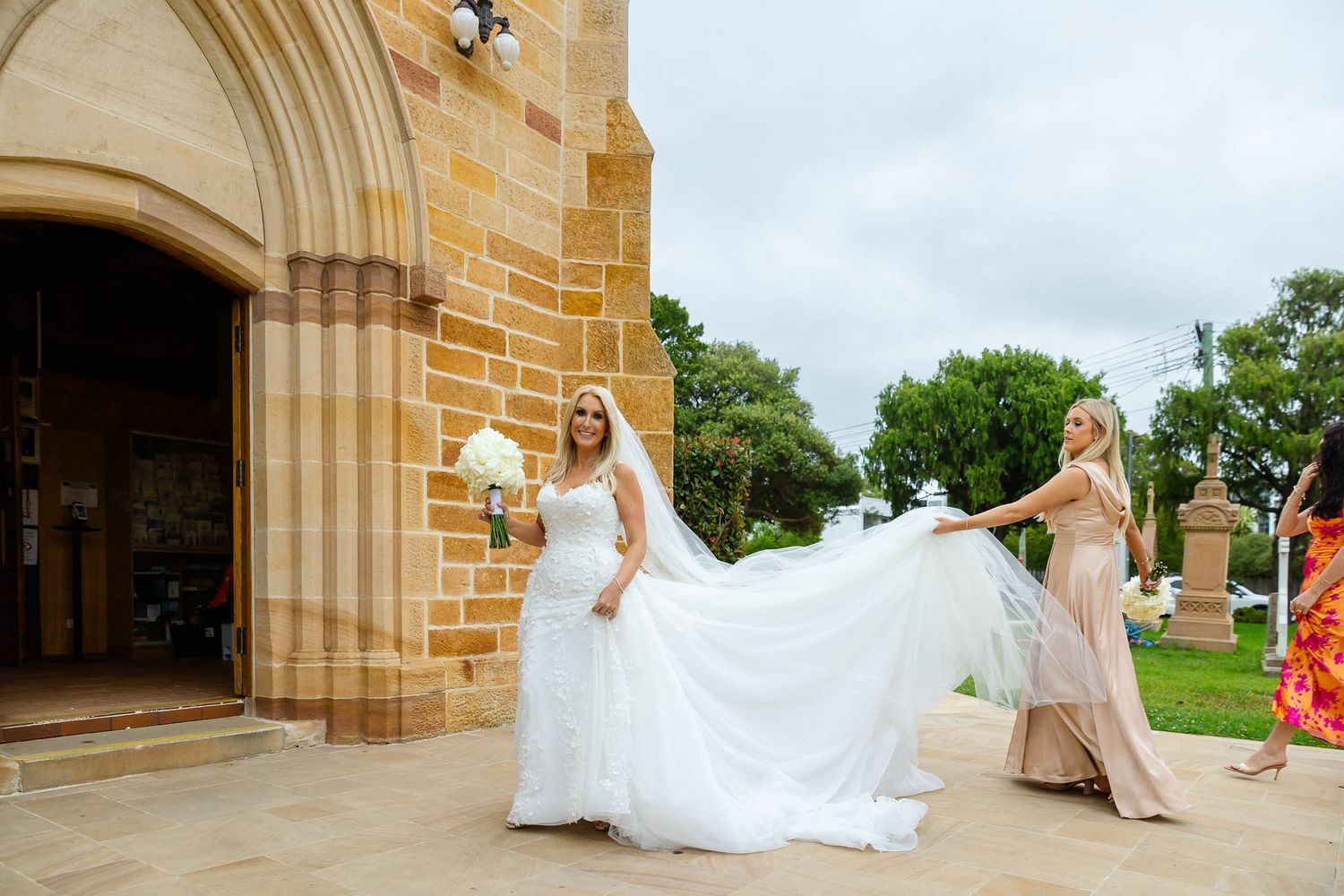 A flowing white wedding dress billows in the wind against a sandstone church exterior.
