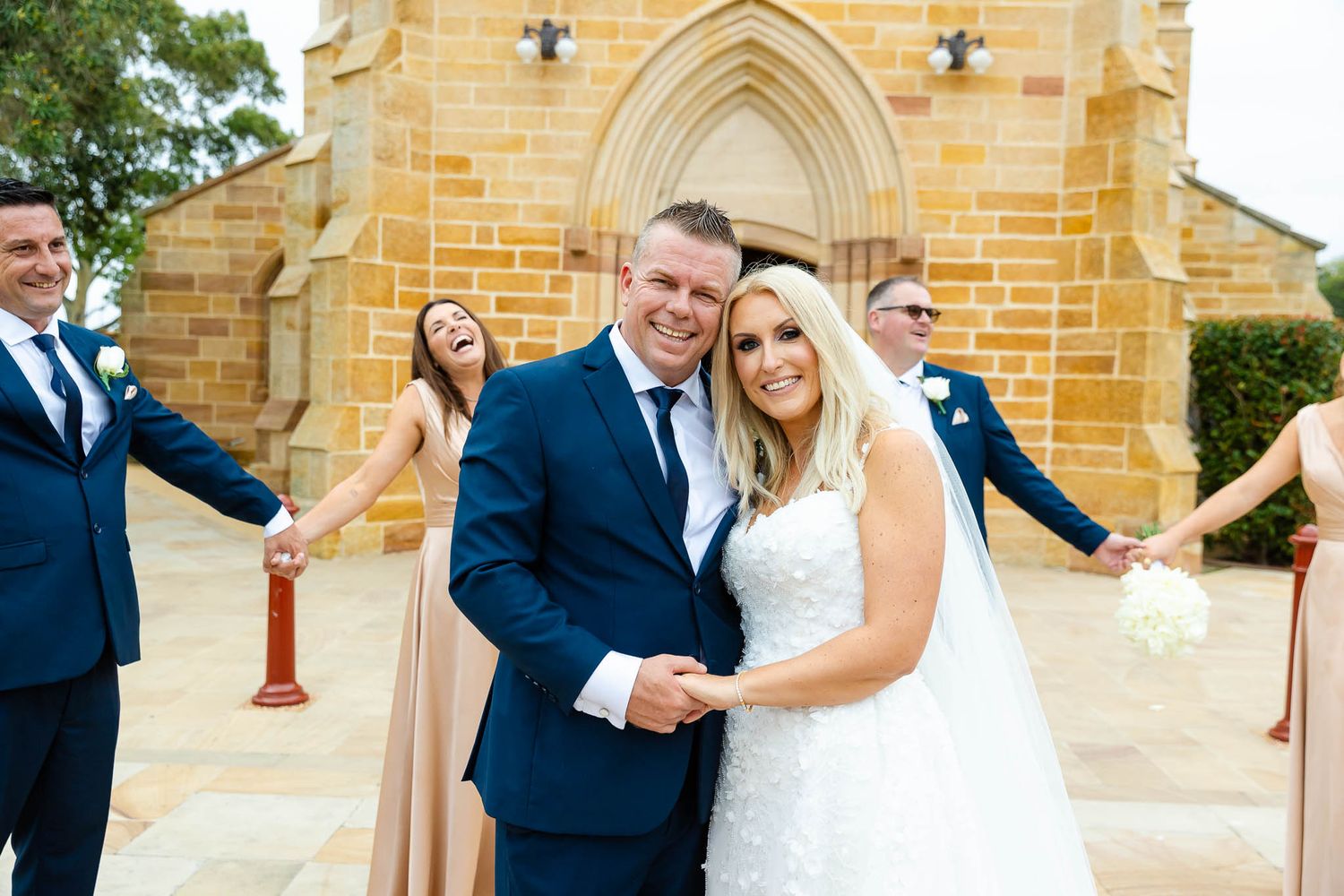 Wedding party members pose in formal attire outside a gothic-style sandstone church.