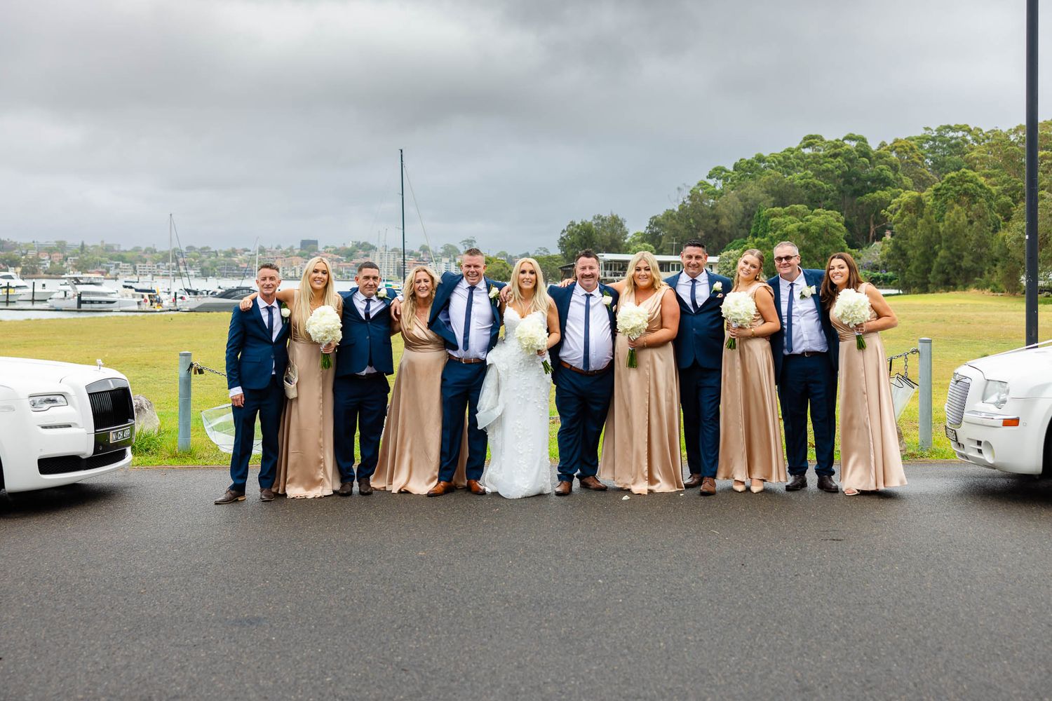 Wedding party members lined up against a scenic waterfront backdrop with boats and buildings visible in the distance.
