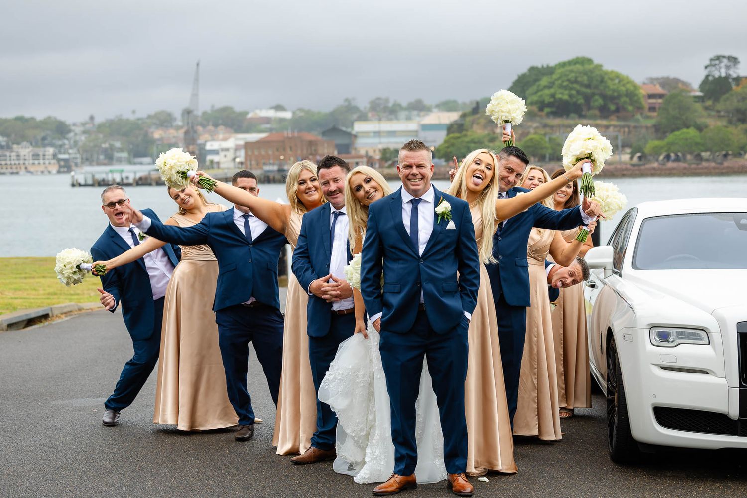 Wedding party in navy suits and beige dresses pose playfully together outdoors near a white car.