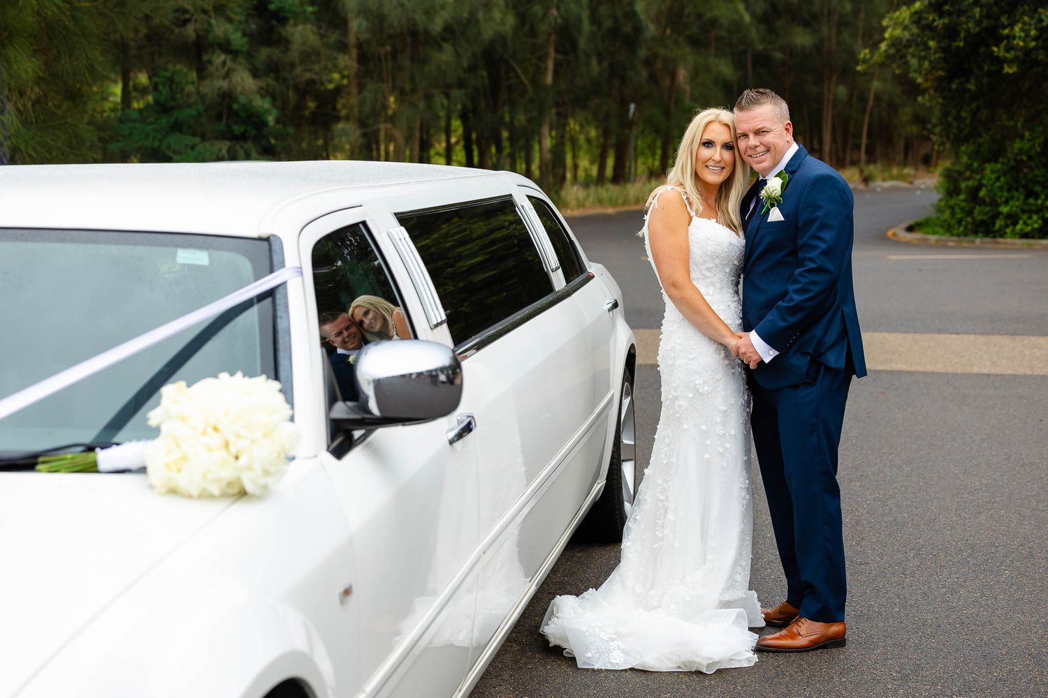 A white stretch limousine parked on a sunny day with people gathered around it for a wedding celebration.