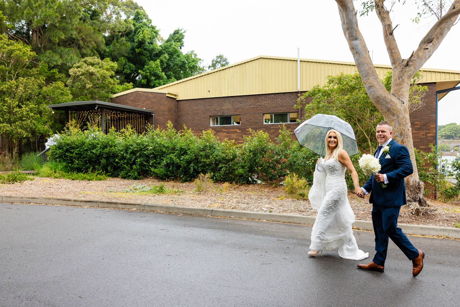 Two people walk down a rainy street with umbrellas outside a brown building with trees.