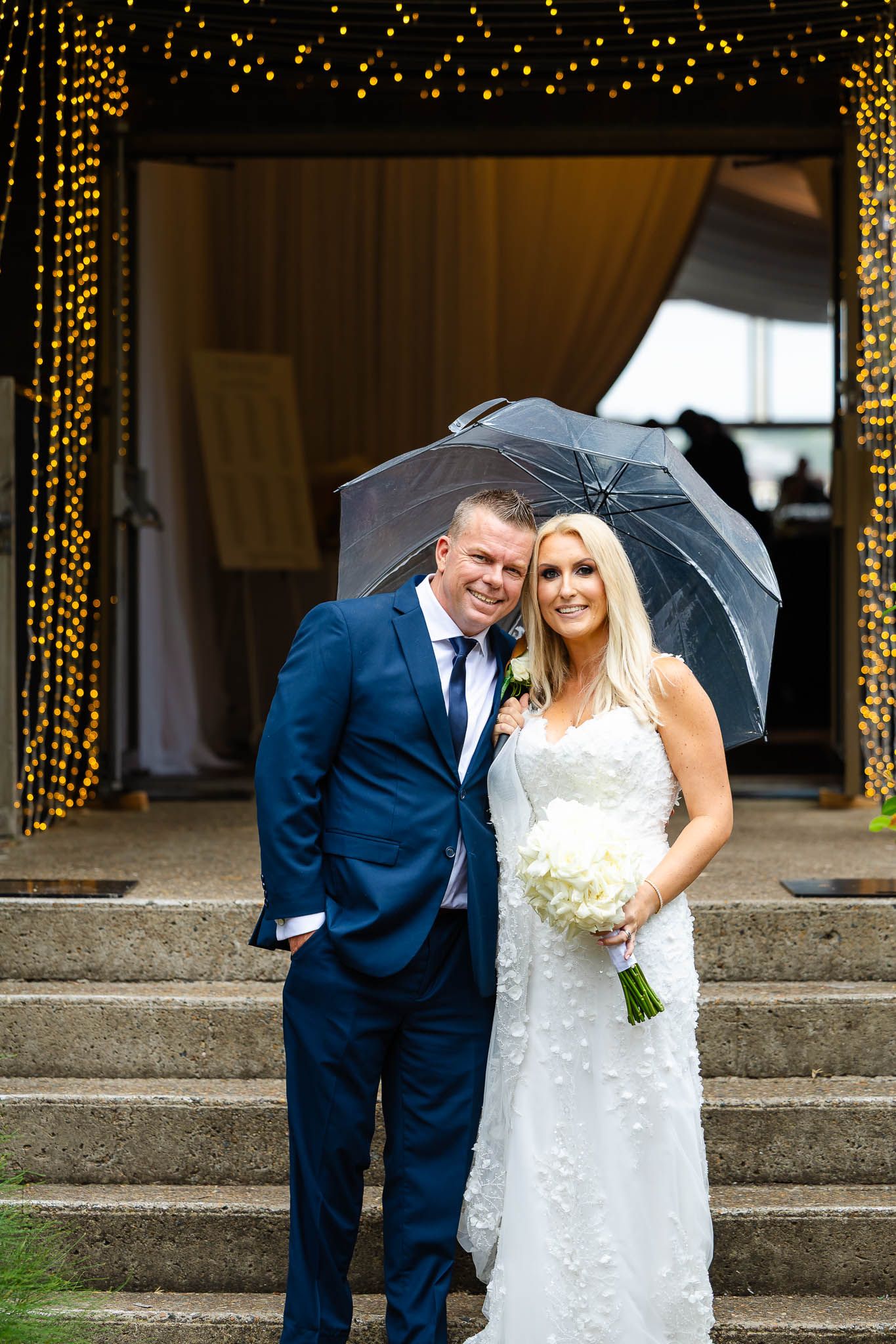 A couple in wedding attire stands under a clear umbrella on stone steps with twinkling lights in the background.