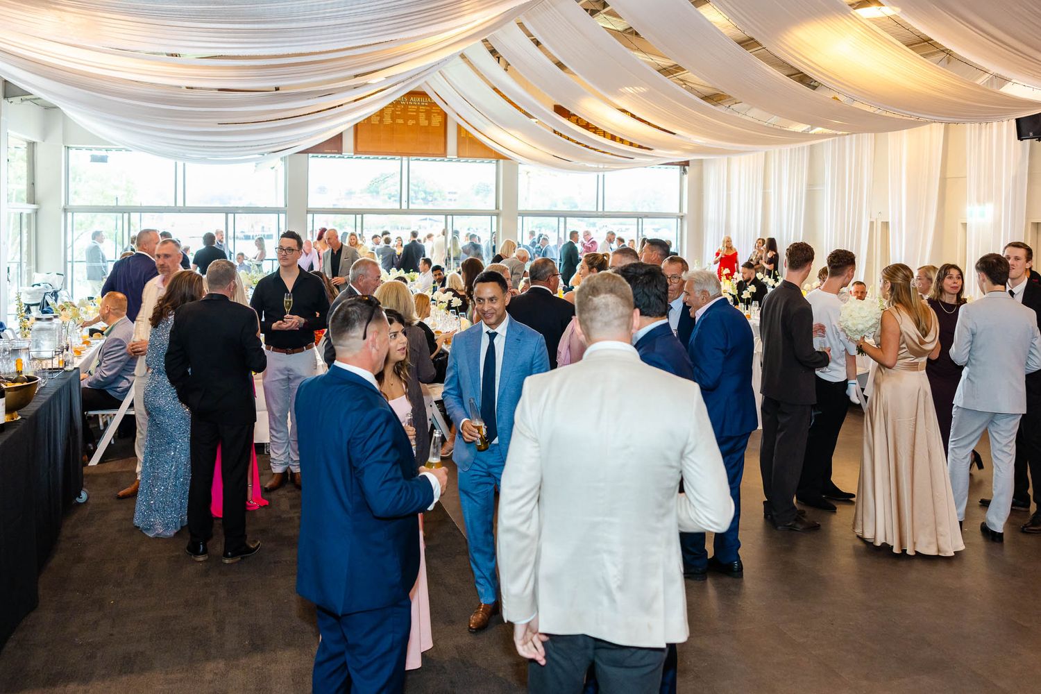 Wedding guests mingle and dance under elegant white drapes at an indoor reception venue.