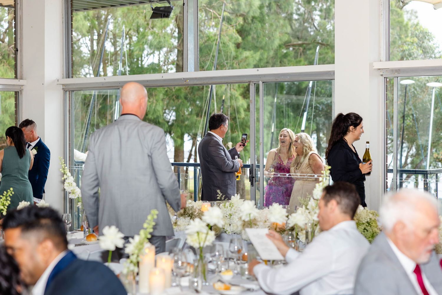 Wedding reception guests seated at elegant tables in a bright sunlit venue with floral centerpieces.