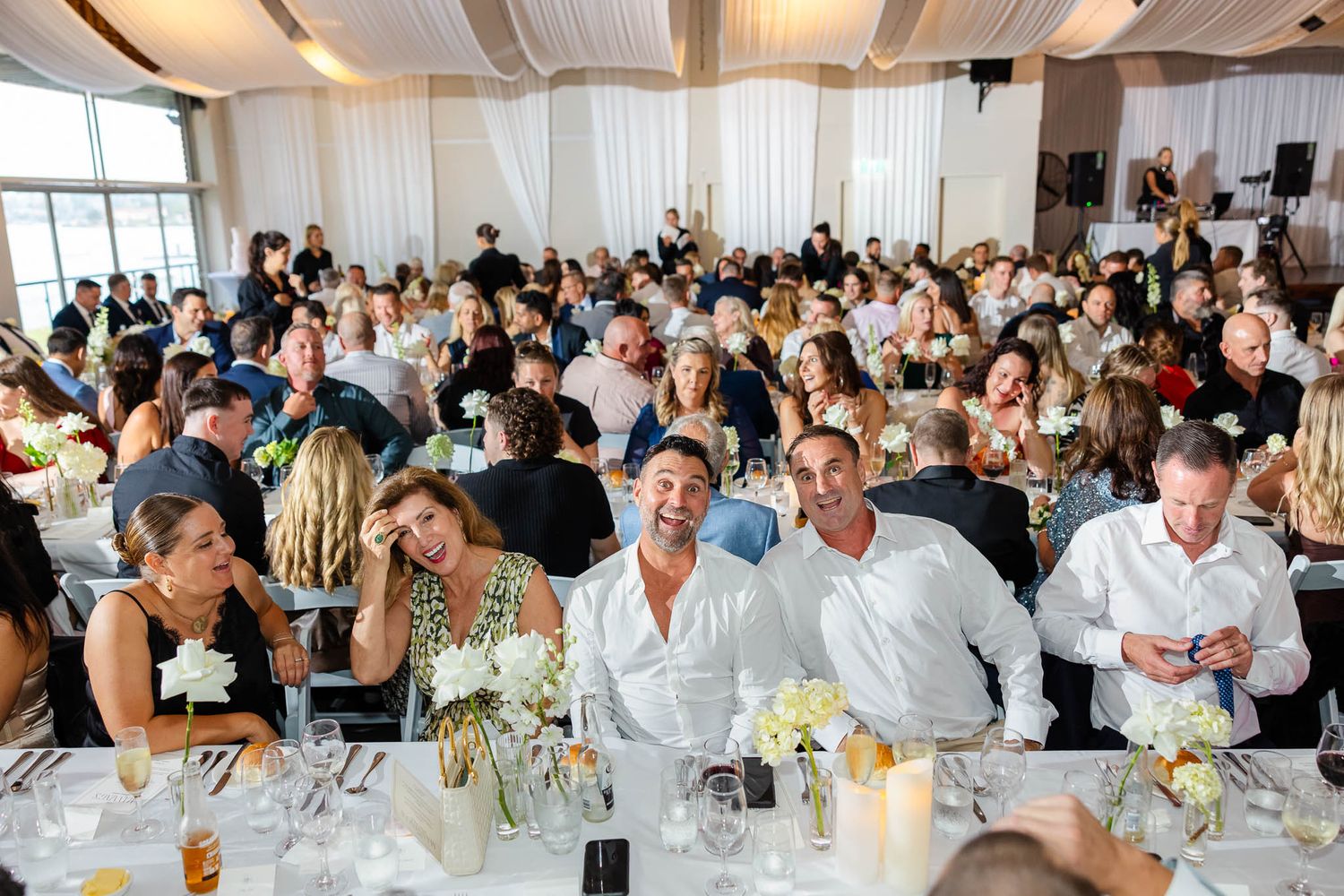 Wedding reception guests seated at long tables in a bright white room filled with natural light.