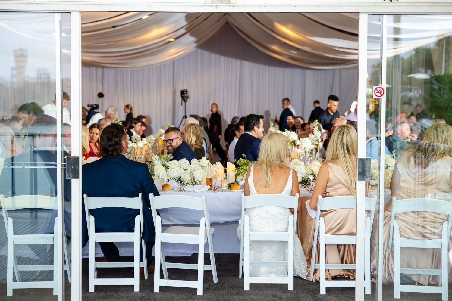Wedding reception guests seated at elegant white tables with draped fabric decor and floral centerpieces.