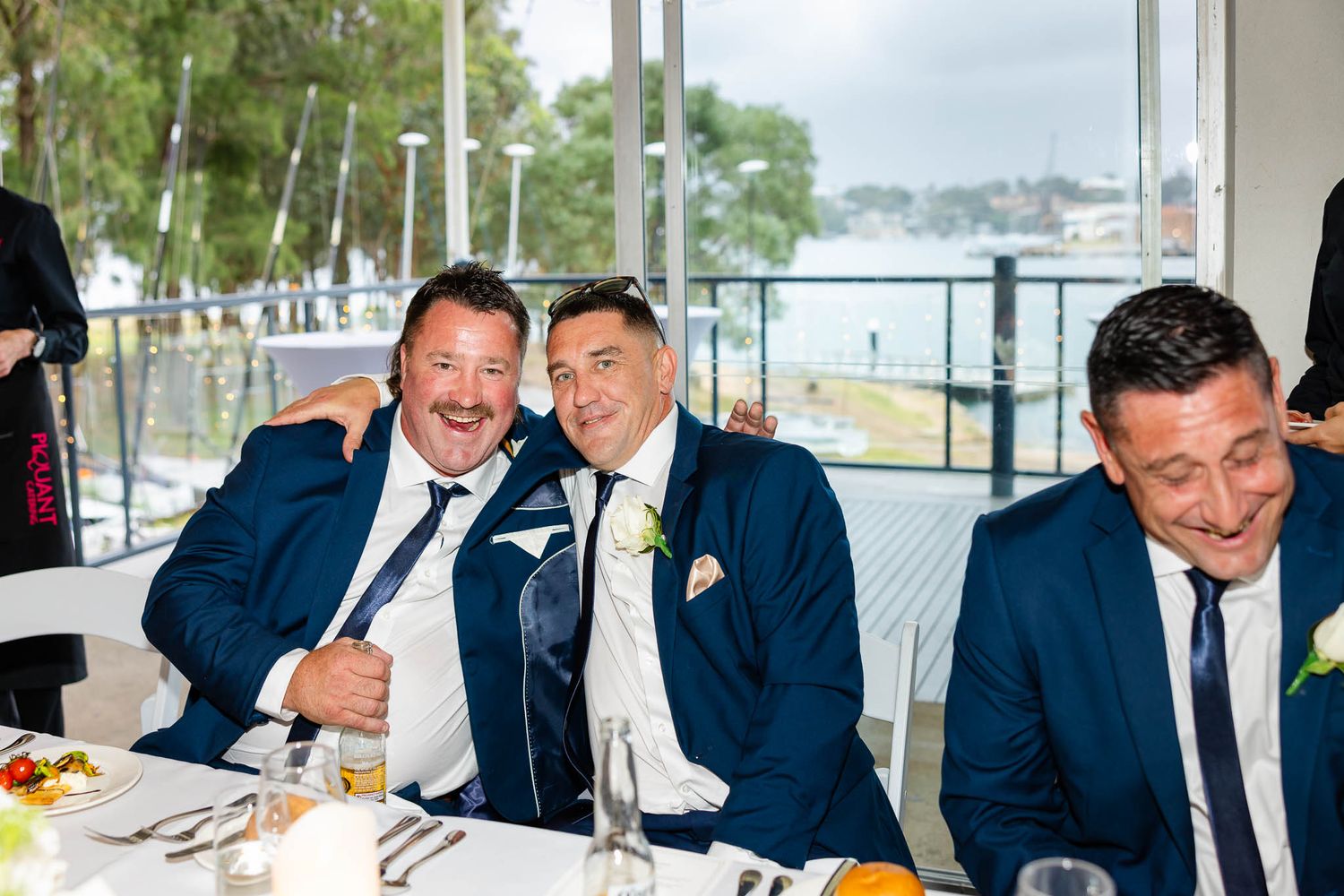 Wedding guests in navy suits share laughter and conversation during a reception dinner at a waterfront venue.