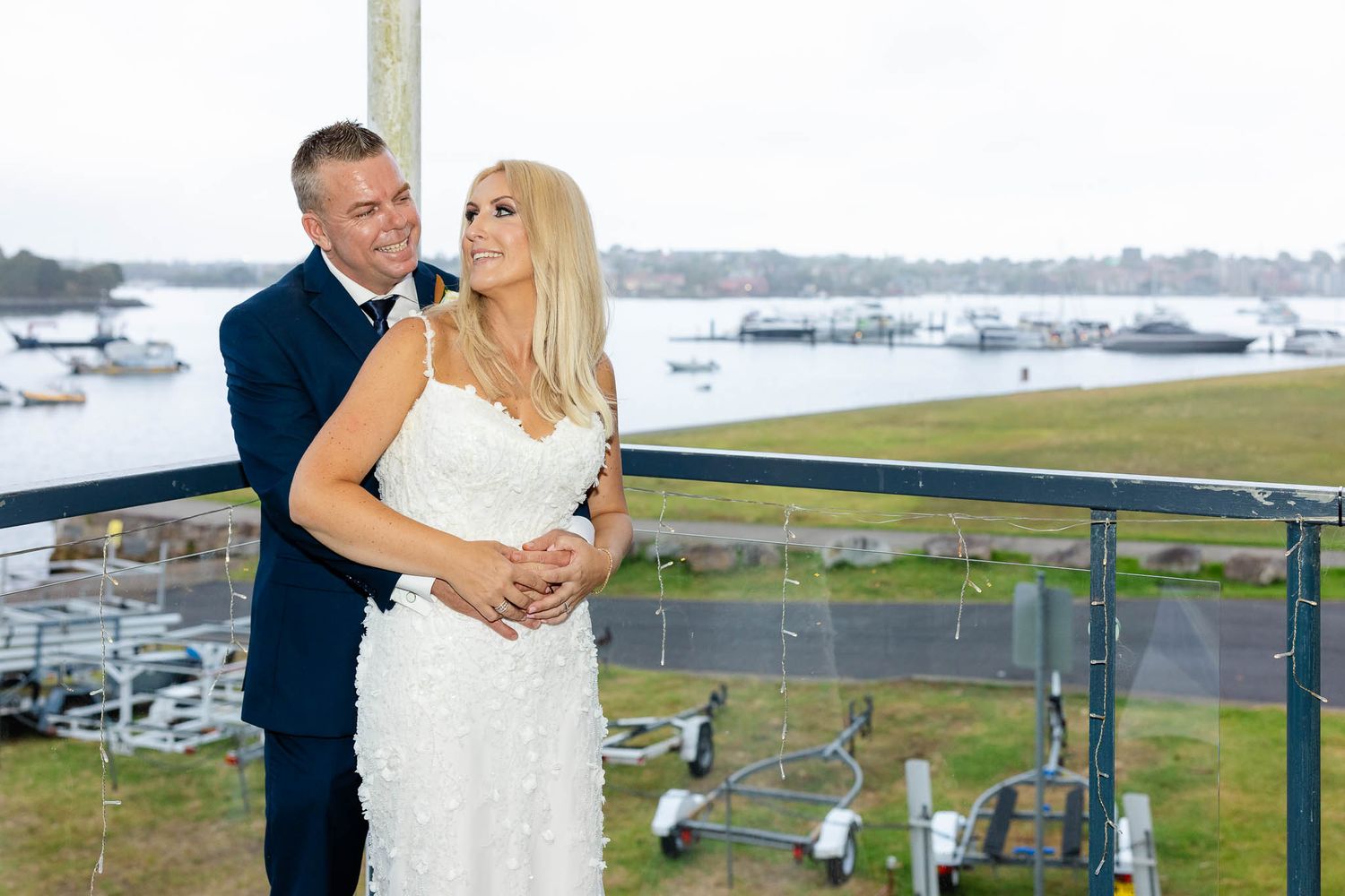 A couple embraces on a balcony overlooking a marina and grassy waterfront area on their wedding day.