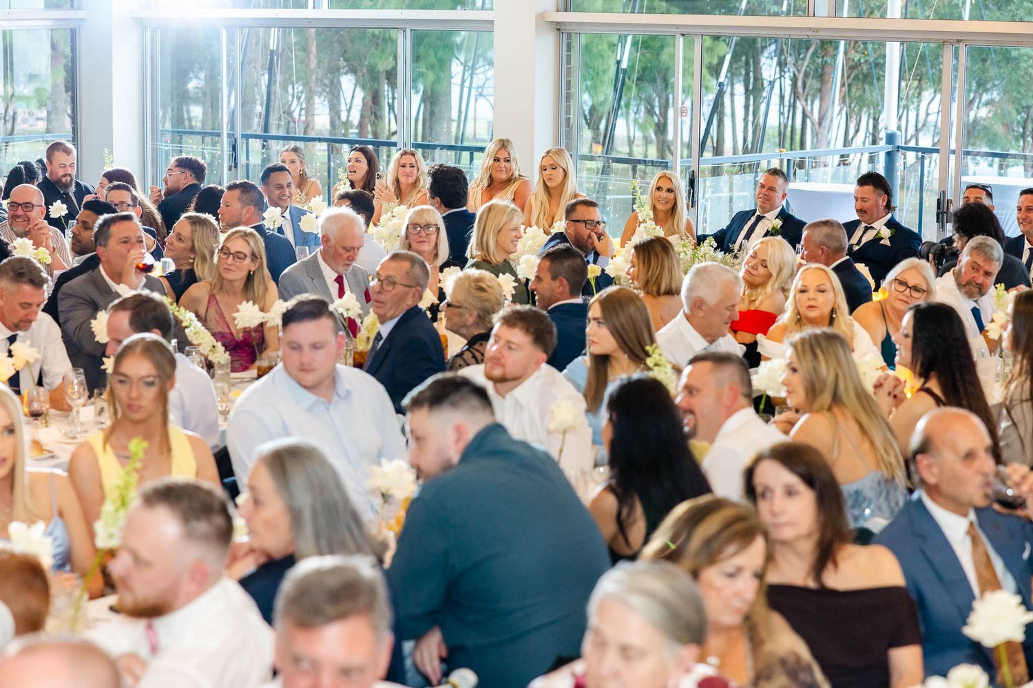 A large group of wedding guests celebrating at an elegant reception venue with glass walls and natural lighting.