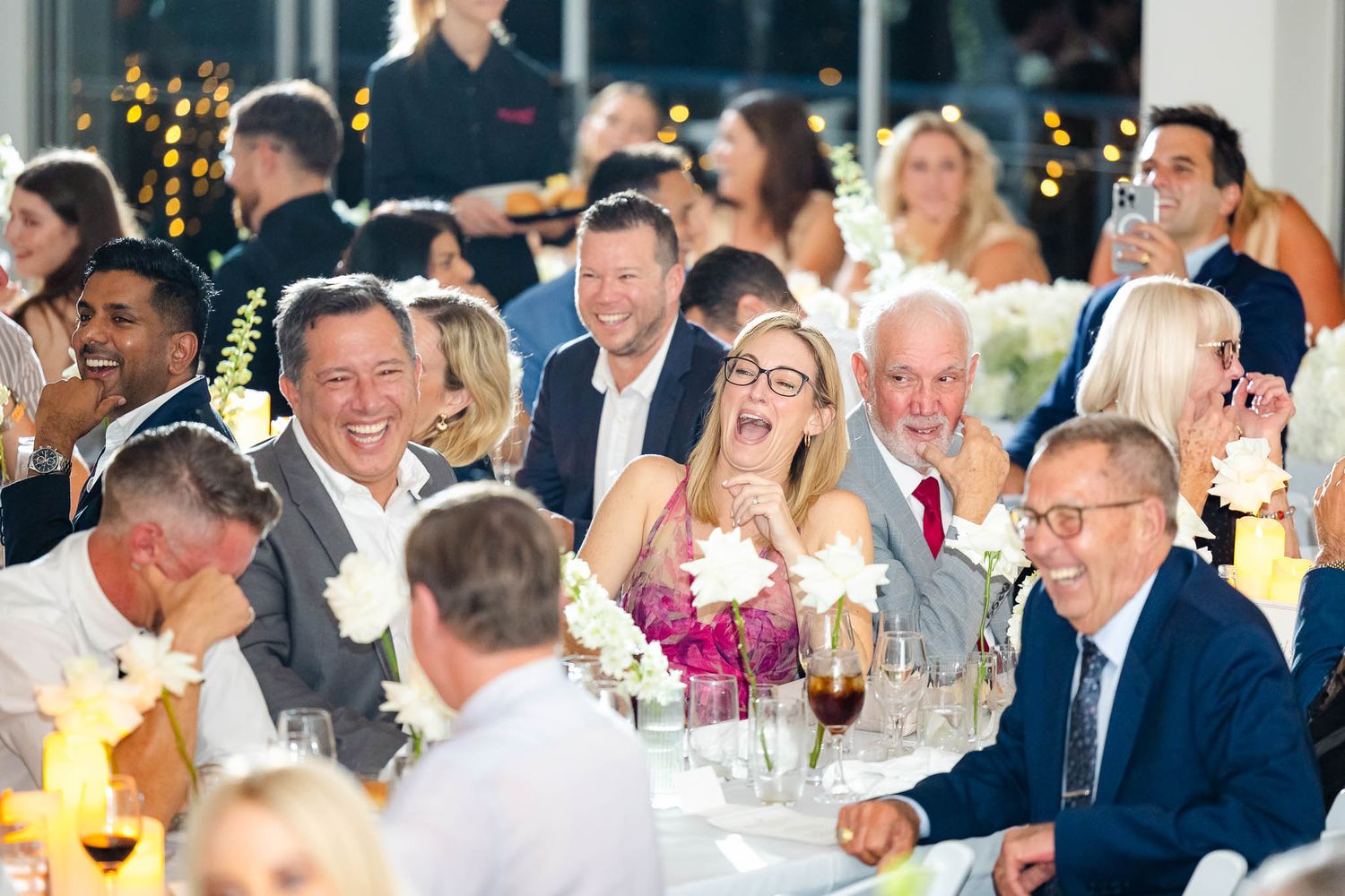 Wedding guests laugh and enjoy themselves at a reception dinner table with glowing candlelight and festive atmosphere.