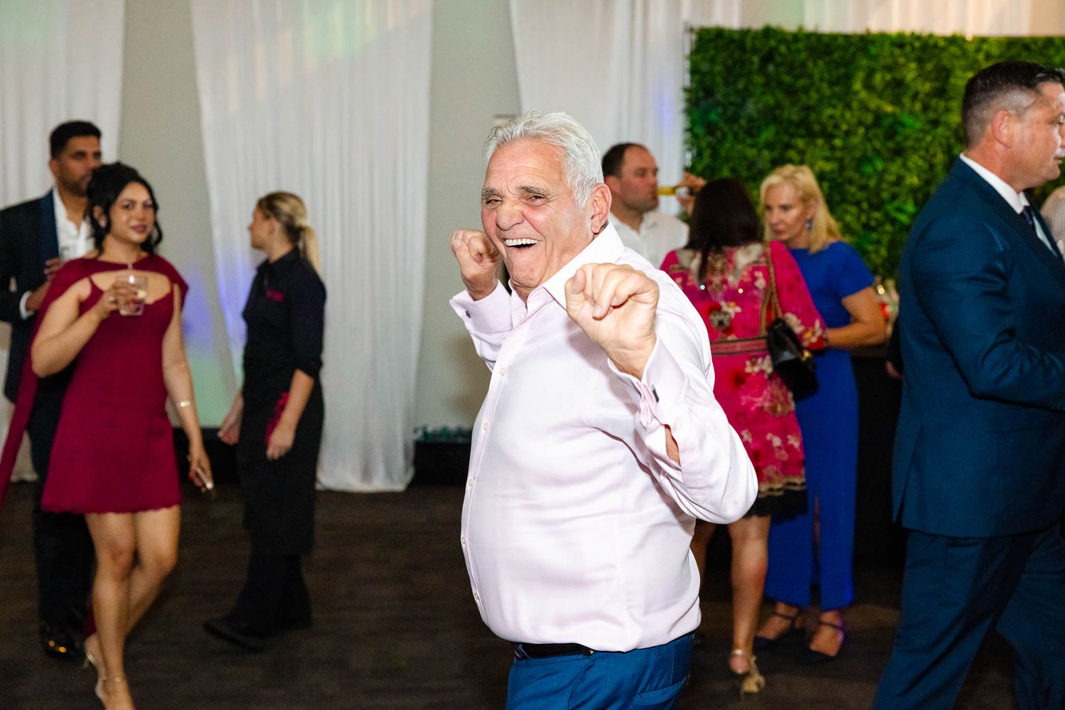 Wedding guests dance and celebrate together on a reception dance floor with white drapes and greenery in the background.