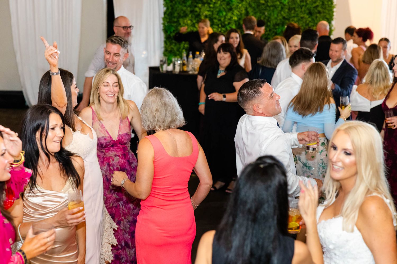 Wedding guests dance and celebrate together on a crowded dance floor at an indoor reception venue.