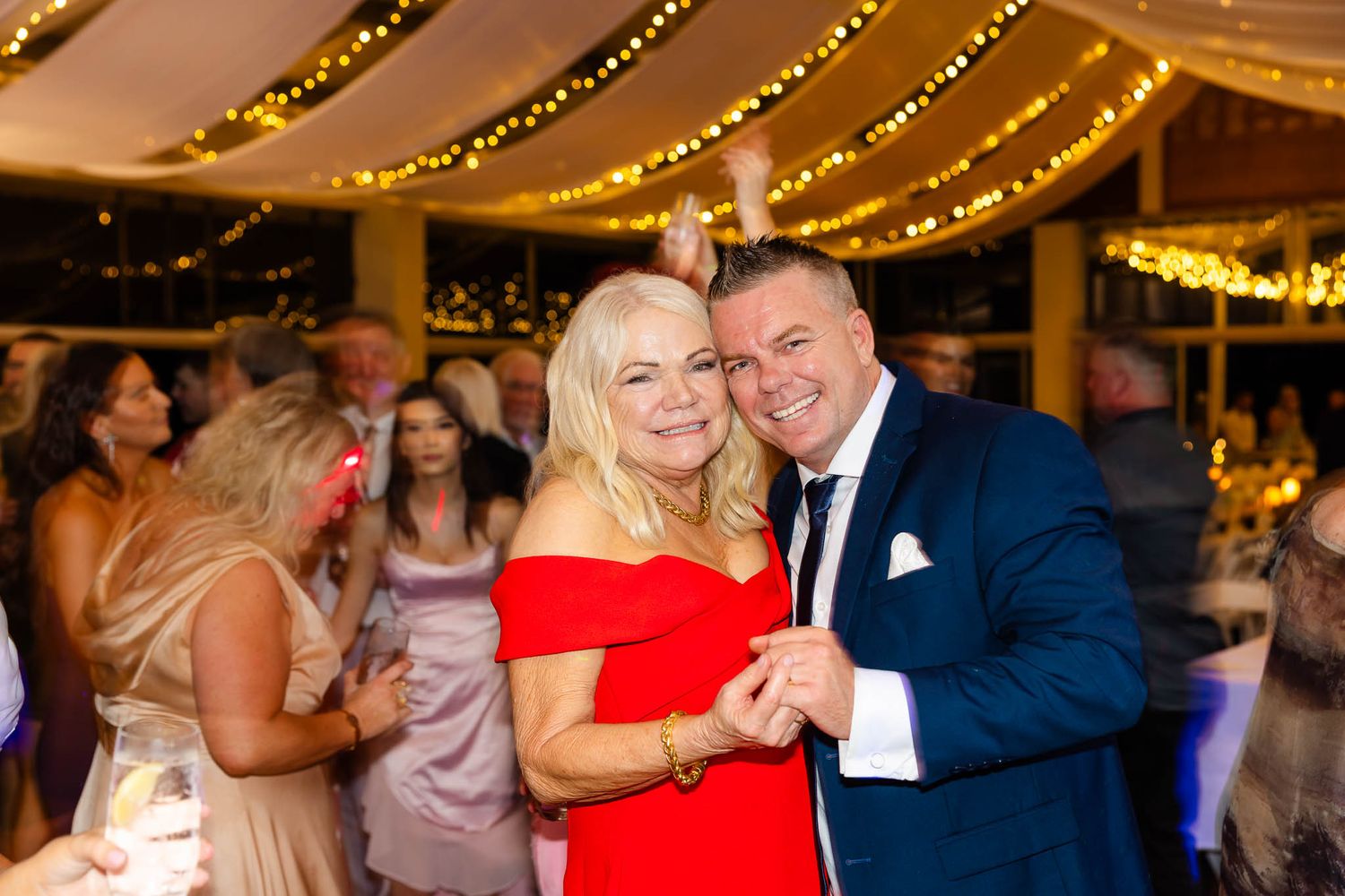Groom and his mother dancing together under romantic string lights at an elegant evening celebration.